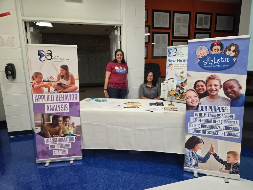 Two people sit behind a table at an event. The table has brochures and decorations. Behind them are banners for Beyond Expectations and Lotus Learning & Arts Center, featuring information about applied behavior analysis and individualized education.