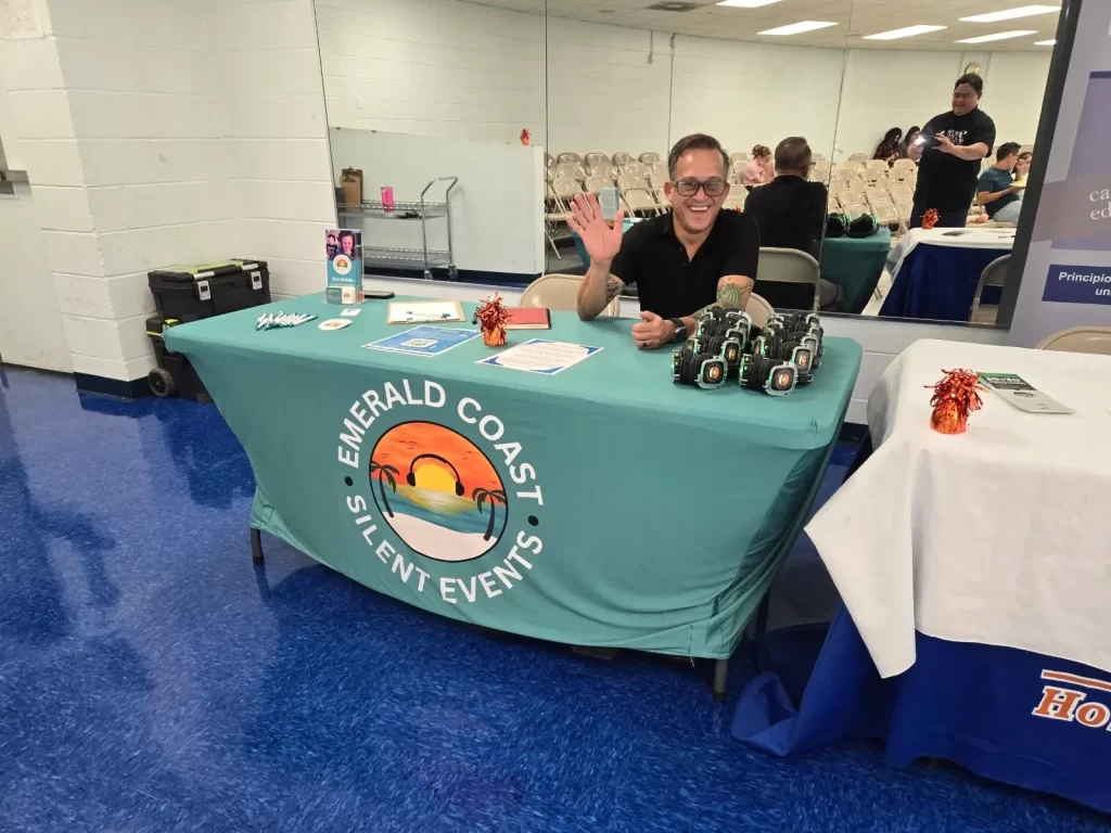 A person sitting at an "Emerald Coast Silent Events" booth waves at the camera. The table is covered with a blue cloth featuring the logo and name. Brochures and other materials are displayed on the table in a room with blue flooring.