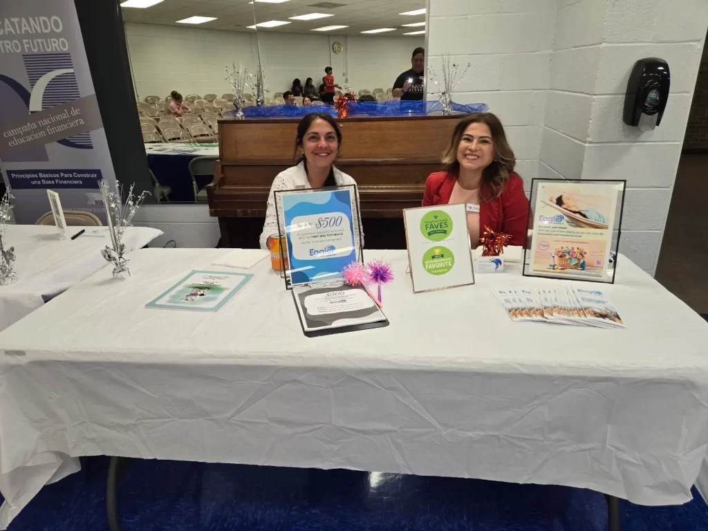 Two women are seated at a table covered with a white cloth, displaying brochures and framed certificates. Behind them is a piano. The setting appears to be an indoor event or fair.