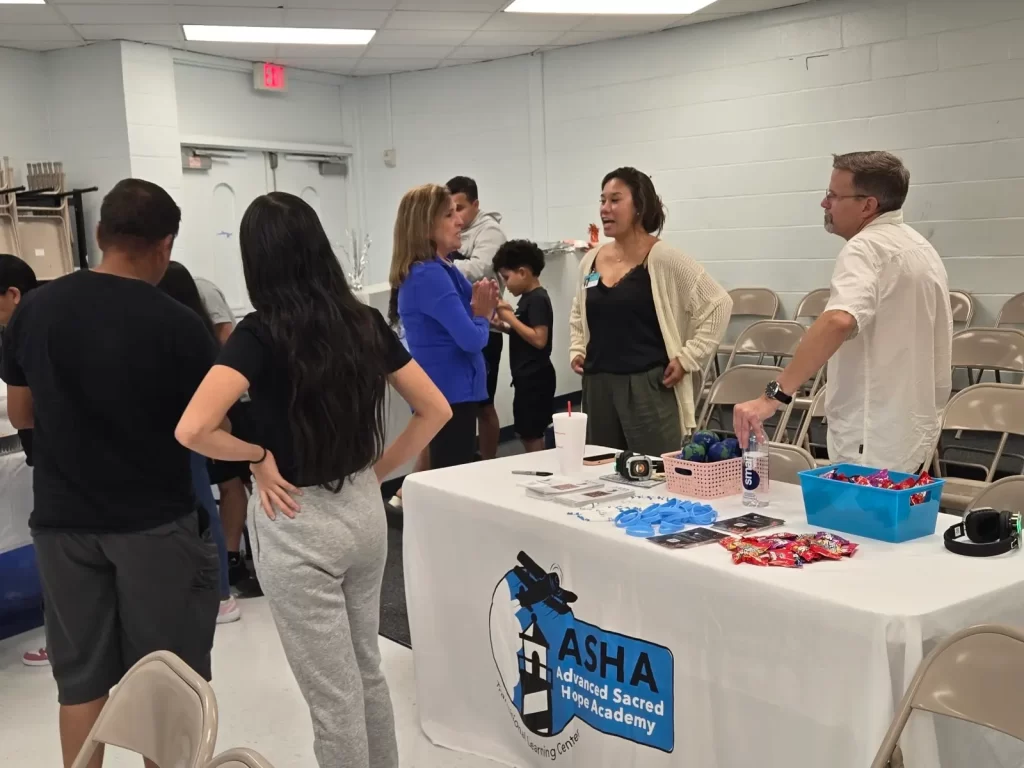 People are engaged in conversation around a table with promotional materials for ASHA Advanced Sacred Heart Academy. The table holds various items, including candies and flyers. Folding chairs are set up in the room with light gray walls.