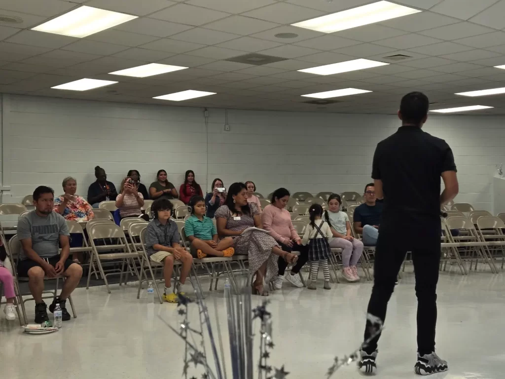 A person stands, addressing a seated audience in a room with fluorescent lighting. The attendees, including adults and children, are seated on folding chairs. The room has white walls and a tiled floor.