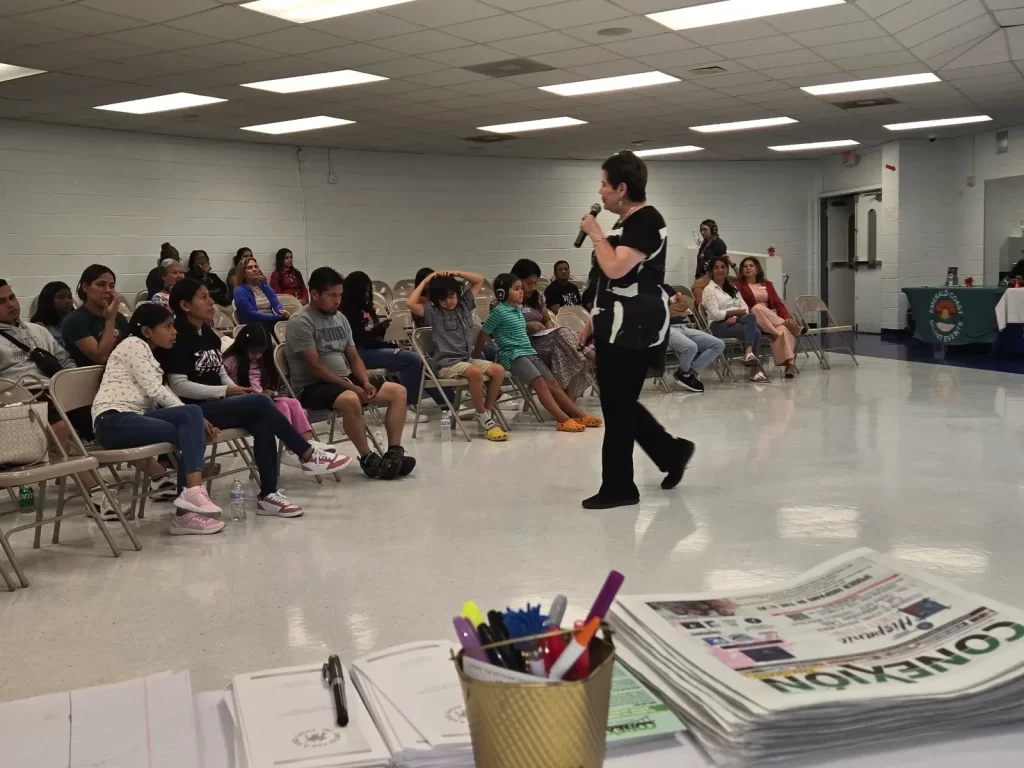 A woman stands speaking into a microphone in a community hall. Seated people listen attentively. In the foreground, there's a table with papers, pamphlets, and a container of pens. The room has fluorescent lighting and neutral-colored walls.