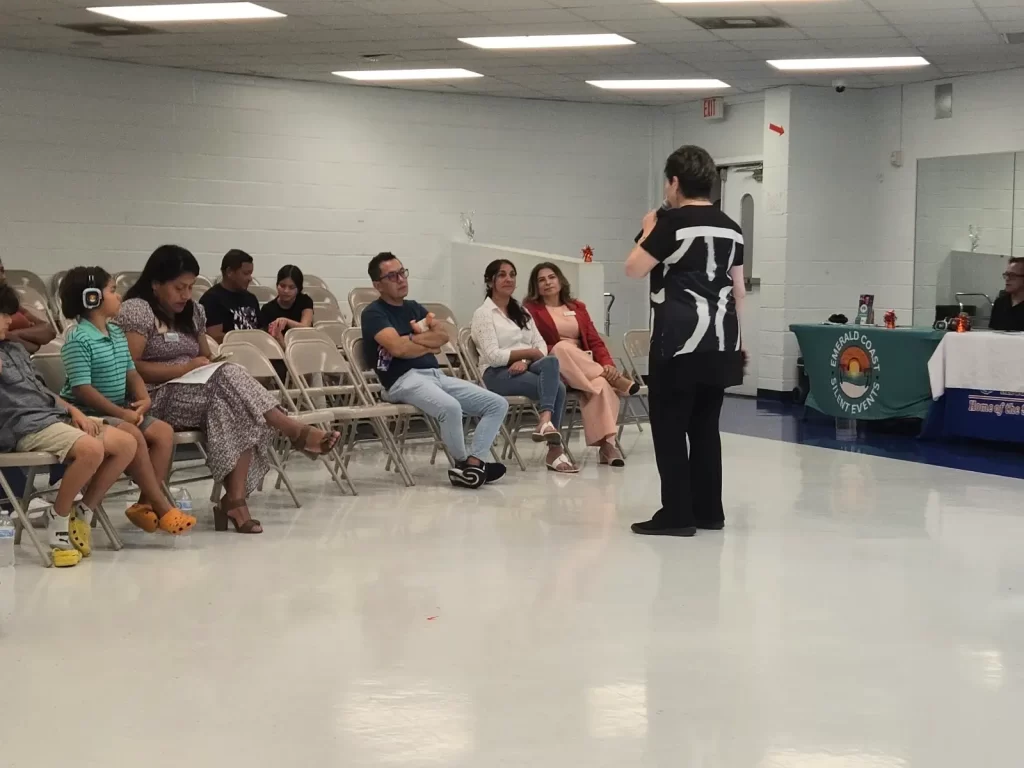 A group of people sit on chairs in a community meeting room. A speaker stands in front of them, gesturing. On the right, a table displays a banner with text and logos. The room is brightly lit with fluorescent lights.