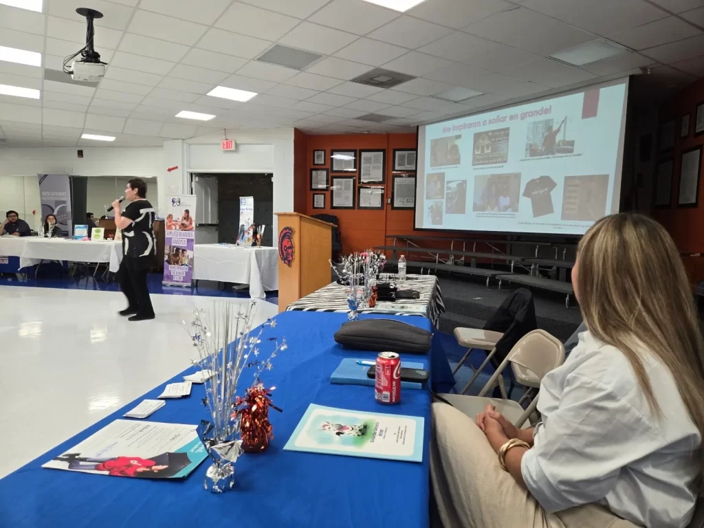 A woman stands and speaks at a presentation in a meeting room, with chairs arranged around tables covered in blue tablecloths. A slideshow is projected on a large screen. Attendees are seated and listening attentively.