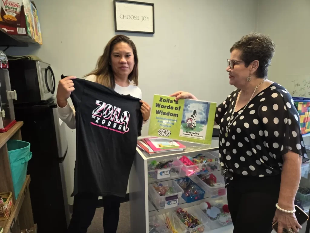 Two women stand in a room. One holds up a black T-shirt with white and pink text. The other holds a book titled "Zoila Words of Wisdom." Shelves with toys and a mini fridge are in the background. A sign above reads "Choose Joy.