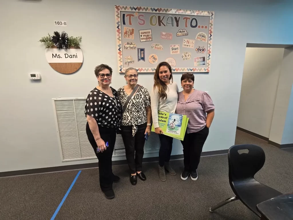 Four women stand smiling in a classroom under a bulletin board titled "It's Okay To..." The board displays various encouraging messages. One woman holds a colorful children's book. A door sign reads "Ms. Dani.