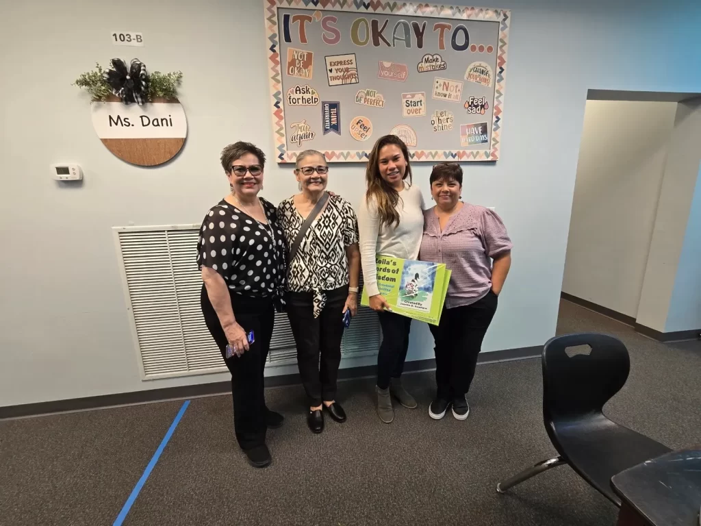 Four women stand together in a classroom, smiling. One holds a brightly colored book. The wall behind them features decorations, including a board that says "It's okay to..." along with various positive affirmations.