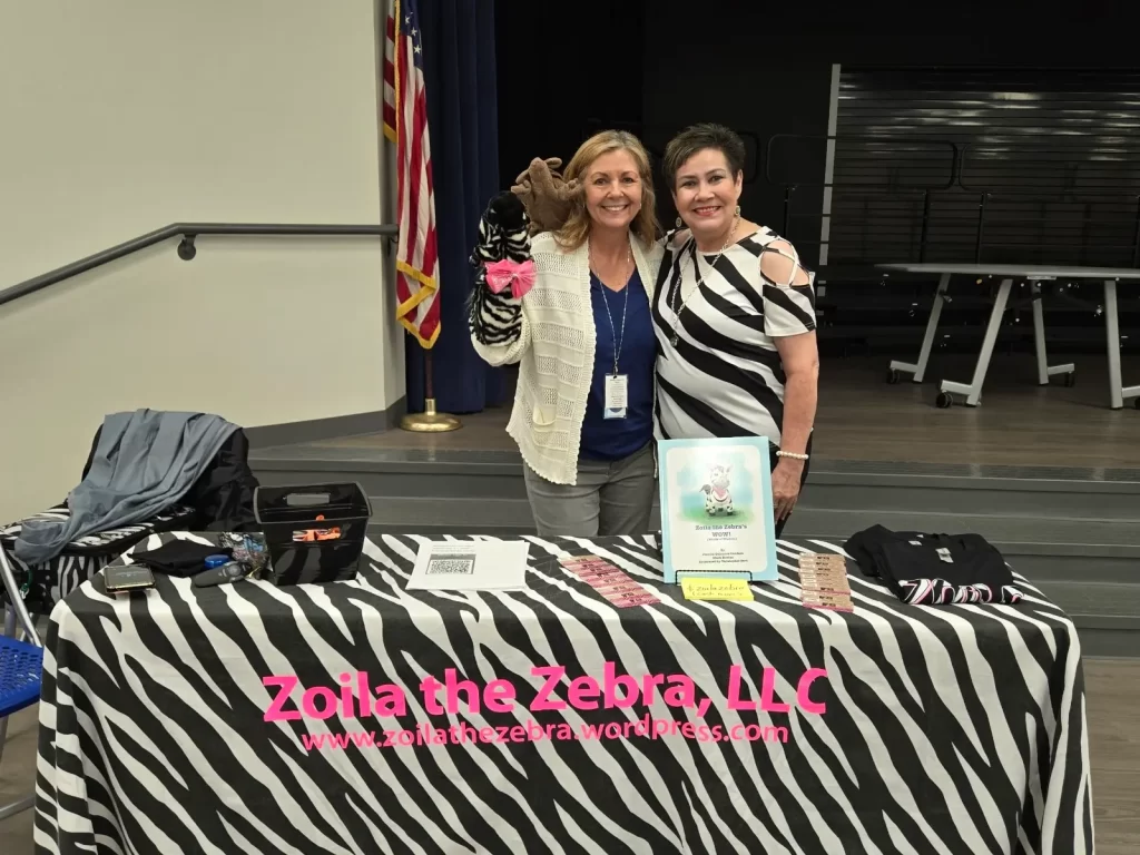 Two women smiling behind a table covered with a zebra-print cloth. The table features a sign that reads "Zoila the Zebra, LLC" and displays brochures and a stuffed zebra puppet. An American flag is in the background.