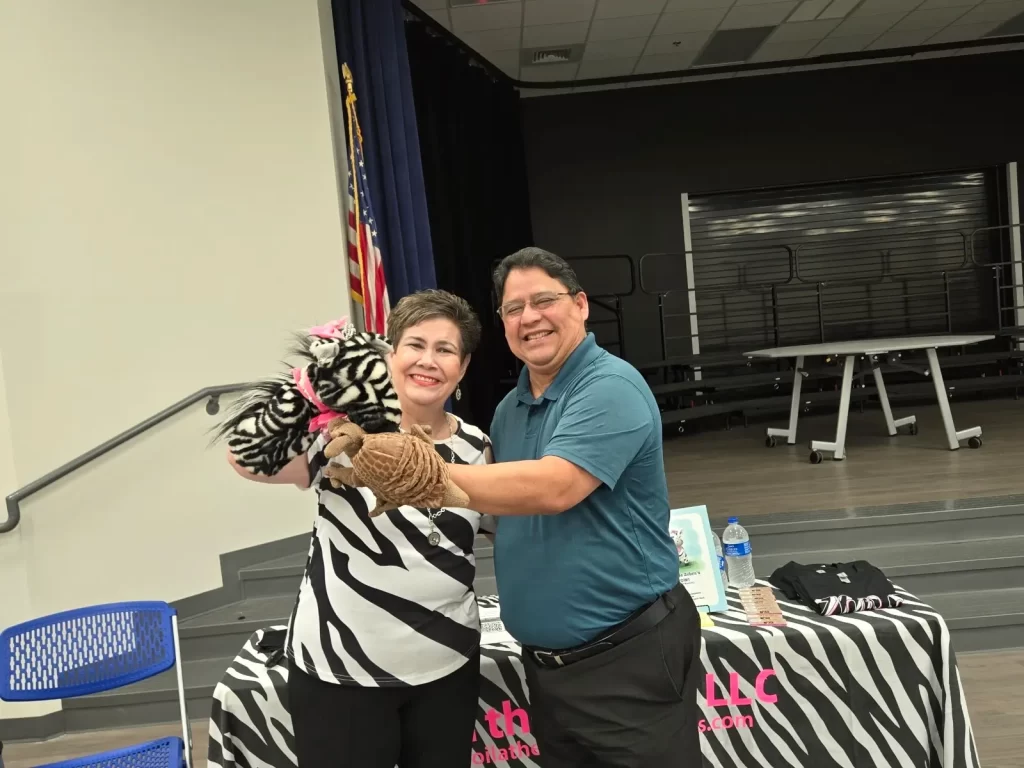 A smiling woman and man stand together indoors, holding animal puppets. The woman holds a zebra puppet, and the man holds a monkey puppet. They are in front of a table with a water bottle and some items. An American flag is visible in the background.