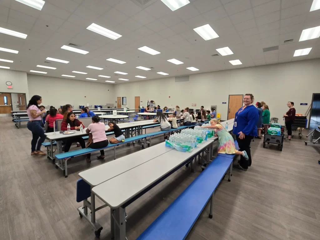 A large room with a group of people sitting at long tables. Some are eating and drinking bottled water. In the foreground, a table holds several cases of bottled water. The room has a high ceiling and neutral-colored walls.