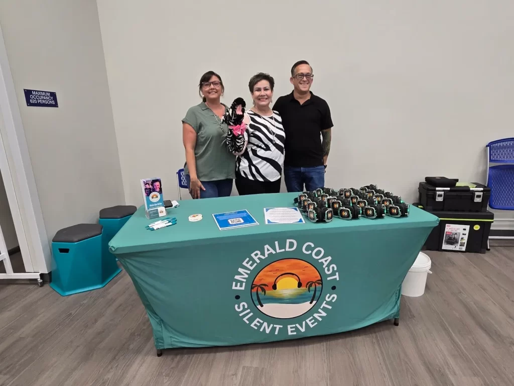 Three people stand behind a table covered with a green tablecloth displaying the "Emerald Coast Silent Events" logo. The table holds various promotional materials and stacks of headphones. The individuals are smiling and posing for the photo.