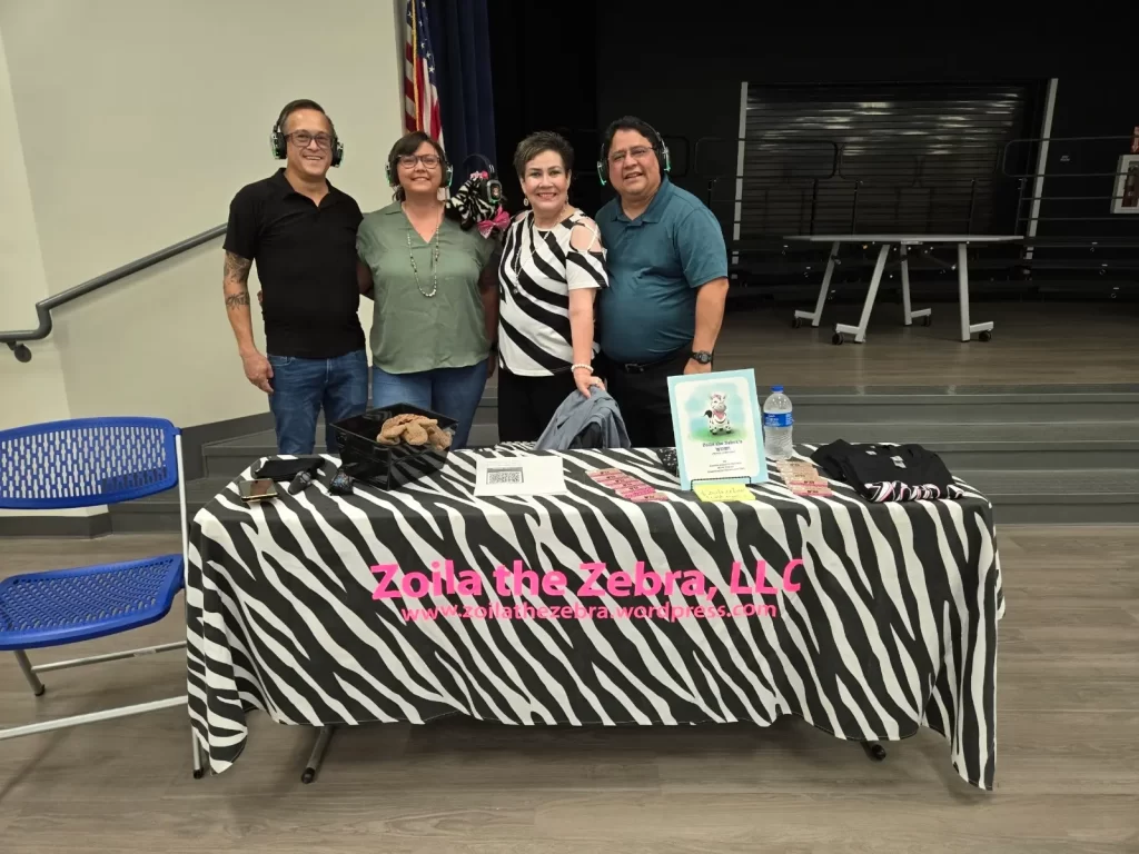 Four people stand behind a table covered with a zebra-striped tablecloth. The table displays promotional materials for "Zoila the Zebra, LLC." An American flag is in the background.