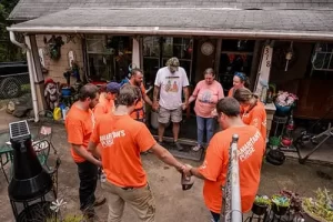 Un grupo de personas con camisetas naranjas se encuentran en círculo cogidas de la mano frente a una casa. Parecen estar rezando o guardando un momento de silencio. La escena se desarrolla al aire libre y hay una variedad de objetos alrededor del porche.