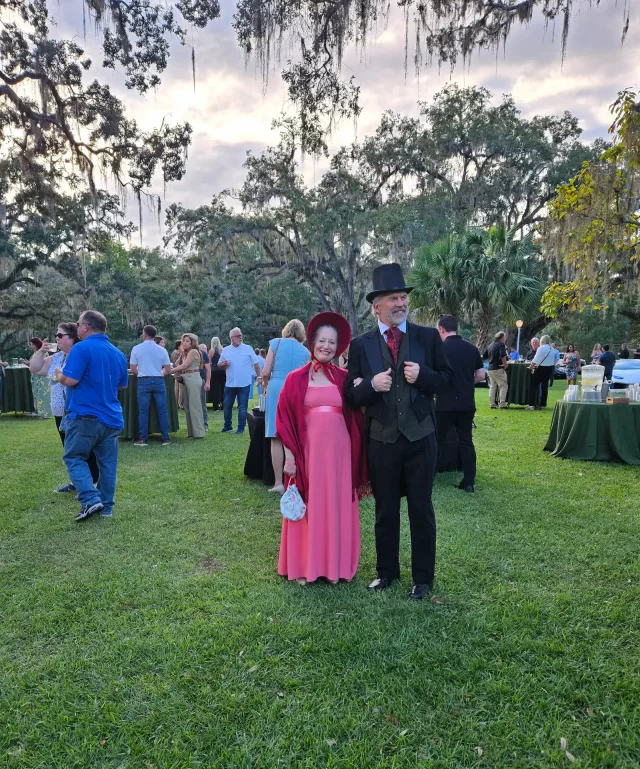 A couple dressed in vintage attire, with the woman in a pink dress and shawl and the man in a black suit and top hat, stand smiling on a grassy lawn. Other people are mingling in the background under large, mossy trees.