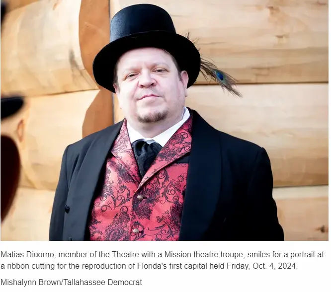 A person wearing a top hat and ornate red vest, with a feather in the hat, smiles for a portrait in front of a log structure.