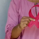 A person wearing a pink blouse holds a pink breast cancer awareness ribbon, emphasizing the importance of early detection and self-exams during Breast Cancer Awareness Month.