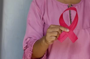 A person wearing a pink blouse holds a pink breast cancer awareness ribbon, emphasizing the importance of early detection and self-exams during Breast Cancer Awareness Month.