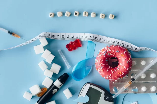 A composition on a light blue background depicting diabetes-related items including sugar cubes, syringes, a measuring tape, a glucometer, insulin pens, a donut with sprinkles, a blue awareness ribbon, and a word "Diabetes" formed with letter beads.