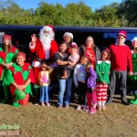 A group of people, including Santa Claus, elves, and children, pose in front of a black limousine outdoors on the Emerald Coast. Some wear festive Christmas outfits, and everyone is smiling. Text reads "Christmas Miracle Children's Fiesta 2015.