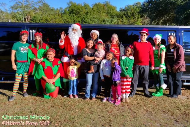 A group of people, including Santa Claus, elves, and children, pose in front of a black limousine outdoors on the Emerald Coast. Some wear festive Christmas outfits, and everyone is smiling. Text reads "Christmas Miracle Children's Fiesta 2015.