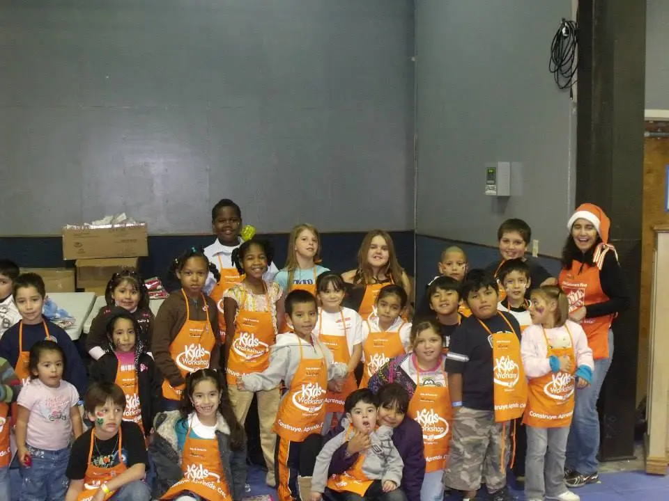 A group of children and adults in orange aprons pose for a photo during a festive kids' workshop, with one person donning a Santa hat, capturing the spirit of a Christmas Miracle. The backdrop features boxes against a gray wall, adding to the vibrant scene.