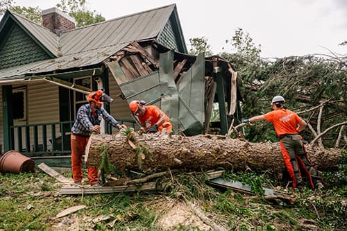 Three workers in safety gear from Samaritan's Purse cut a large fallen tree in front of a damaged house. The tree has collapsed onto the roof, causing significant destruction. Debris is scattered around the scene in Asheville.