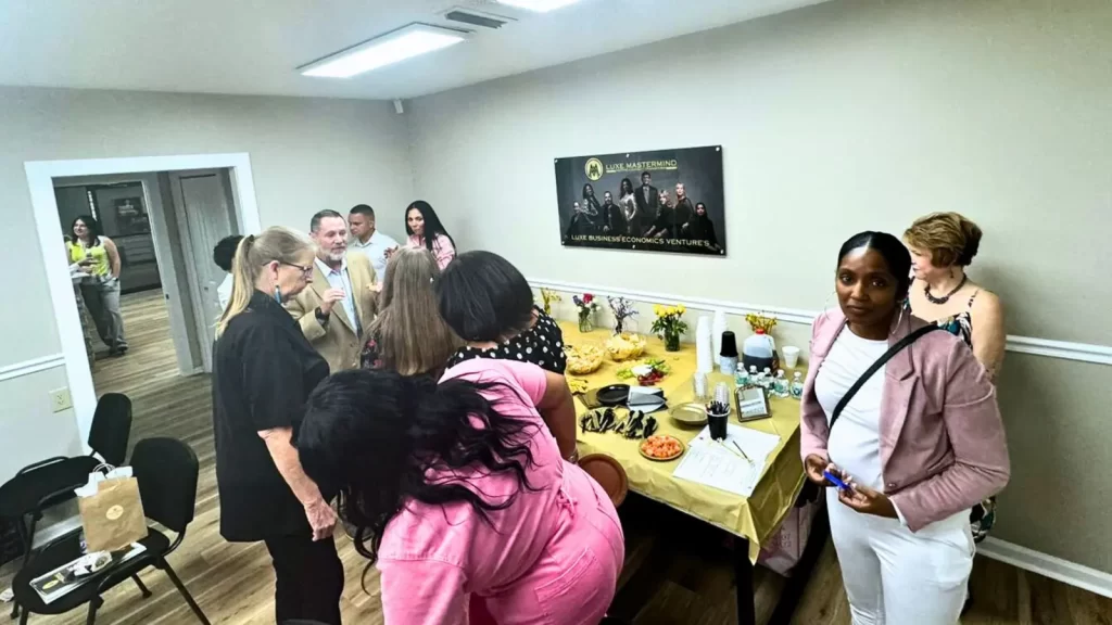 A group of people gather in a room decorated for a celebration. A table is covered with a yellow tablecloth, food, and drinks. Some people stand and chat while others reach for refreshments. In the background, a poster features an award-winning theme.