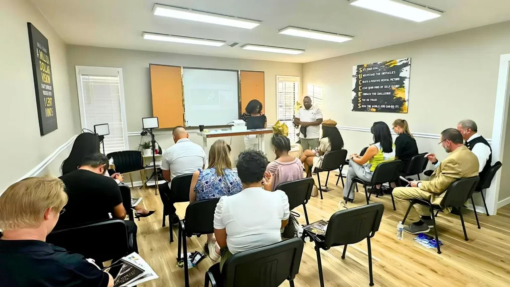 A classroom with seated participants facing a projector screen and two presenters standing at the front. The room is bright with natural and artificial lighting, featuring motivational posters on the walls.