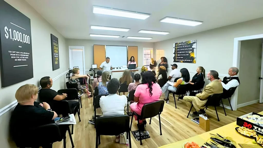 A diverse group of people sits in a well-lit room, listening to a speaker at the front. Posters on the walls feature motivational quotes. Everyone is facing forward, engaged in the presentation. Snacks are visible on a table in the corner.