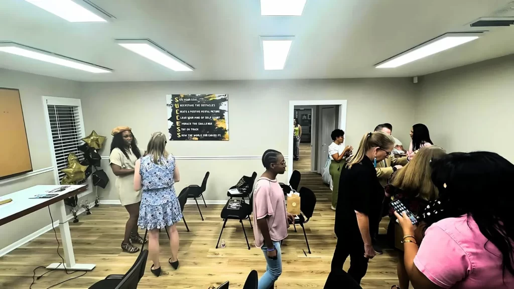 A group of people socializing in a well-lit room with chairs arranged in a semicircle. Some are standing, talking, and checking their phones. A motivational poster is on the wall in the background.