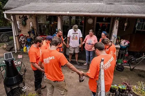 A group of people wearing orange "Samaritan's Purse" shirts stands in a circle, hands clasped in prayer, in front of a house. In the peaceful Asheville area yard adorned with plants and decorations, they reflect on their mission to install water filtration systems for the community.