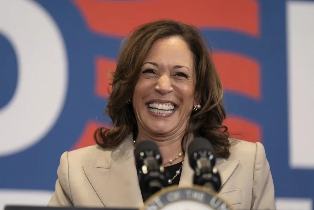 Una mujer con el pelo hasta los hombros, vestida con un blazer beige, sonríe ampliamente. Está de pie frente a un micrófono y un fondo con colores rojo y azul, exudando la energía confiada de Kamala Harris mientras marca el comienzo de una nueva era.