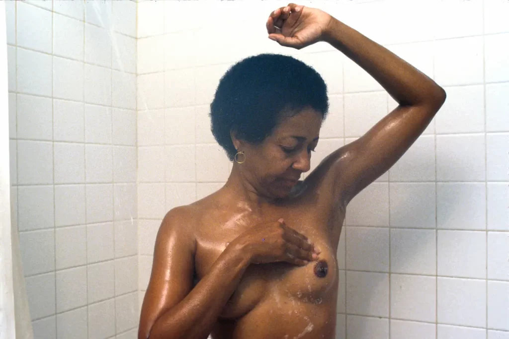 In a tiled shower, water gently cascades down as a person with short curly hair raises one arm, carefully performing a breast self-examination on the right side.