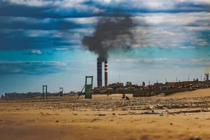 Una playa de arena con escombros dispersos se extiende hacia un pueblo con dos chimeneas que emiten humo oscuro contra un cielo nublado, lo que resalta la necesidad urgente de energía sostenible para cerrar la brecha entre la naturaleza y la contaminación industrial.