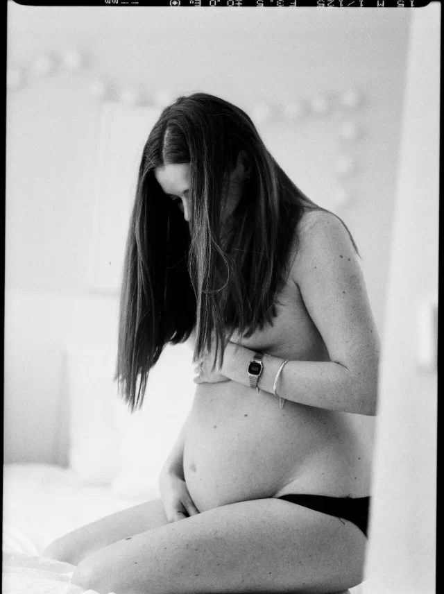 Una mujer embarazada de pelo largo está sentada en una cama adornada con sábanas suaves. Está parcialmente cubierta, sosteniendo su vientre con la mano y mirando hacia abajo con aire pensativo. La imagen en blanco y negro crea una atmósfera serena e íntima.