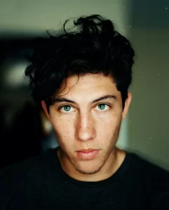 A person with black hair and green eyes looks directly at the camera. Their expression is neutral, and they're wearing a dark shirt. The softly blurred background hints at a scene from Puerto Rico, adding an intriguing layer to their captivating presence.