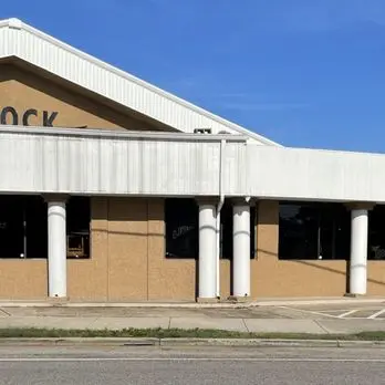 A closed, single-story building with large white columns in front, resembling an iconic landmark on The Block. Featuring boarded windows and a large overhang, the tan exterior contrasts with the clear sky. Partial rooftop lettering is obscured, while a sidewalk and street lie before it.