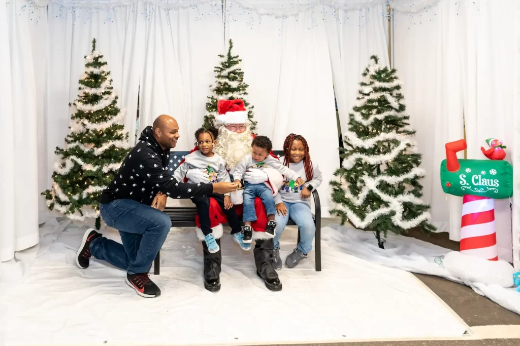 A family poses with Santa Claus in front of a festive backdrop featuring Christmas trees. A man kneels beside two children who sit on a bench with Santa. A decorative sign reading "S. Claus" is visible on the right.