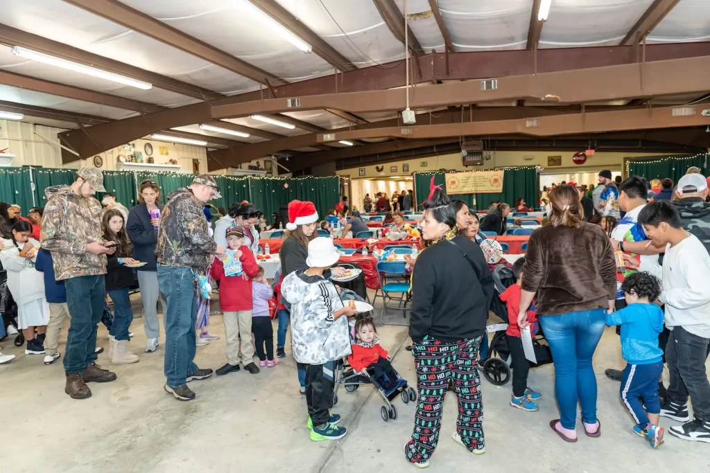 A festive gathering in a large hall, with people of all ages mingling. Some are dressed in holiday attire, like Santa hats. Tables are decorated and filled with food, and the atmosphere is lively and cheerful.