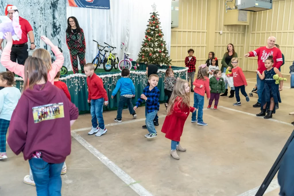 Children and adults are joyfully dancing in a festive indoor setting. In the background, there's a Christmas tree and a few bicycles displayed on a stage. The room is decorated with string lights, adding to the celebratory atmosphere.