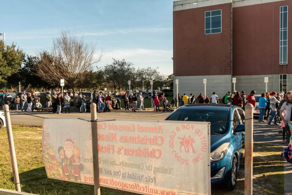 A community Christmas event with people gathered outside a building next to parked cars. A banner in the foreground promotes a Christmas Miracle Children’s Feast. Trees and a blue sky are visible in the background.