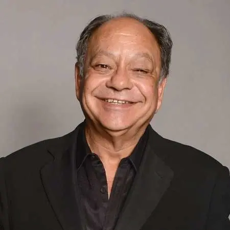 Smiling comedian in a black suit and shirt against a neutral gray background, reminiscent of Cheech Marin's effortless charm.