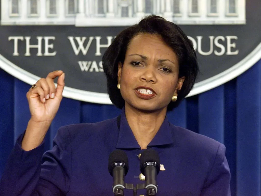 A person in a dark suit, reminiscent of Condoleezza Rice's poised presence, speaks at a podium with two microphones. They are gesturing with their right hand. A sign in the background reads "The White House, Washington.