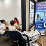 Three people sit at a table in an office discussing health insurance. A large sign on the wall promotes Florida Blue health coverage. Bags of groceries are on a cart nearby.