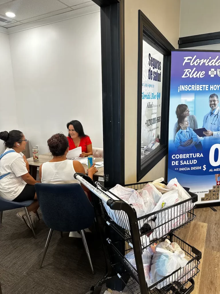 Three people sit at a table in an office discussing health insurance. A large sign on the wall promotes Florida Blue health coverage. Bags of groceries are on a cart nearby.