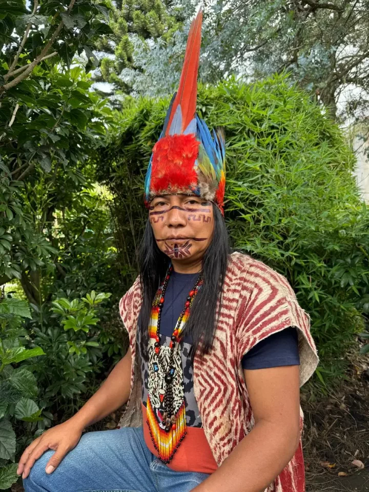 Person wearing a colorful feathered headdress, traditional clothing with patterns, and beaded necklaces. The individual has painted face designs and is sitting outdoors, surrounded by lush greenery.