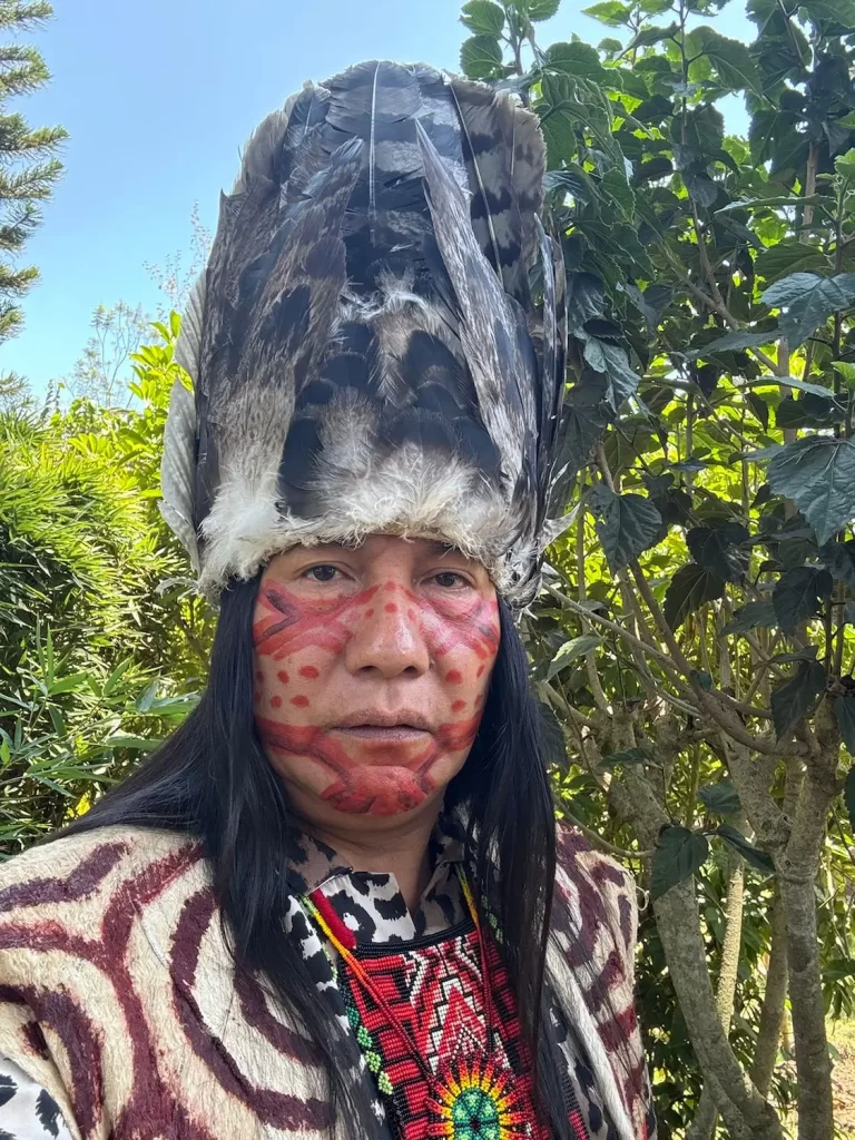 Una persona vestida con un atuendo tradicional, con un tocado de plumas, pintura facial elaborada y un collar de cuentas, se encuentra al aire libre. Está rodeada de una vegetación exuberante y la luz del sol se filtra a través de las hojas.