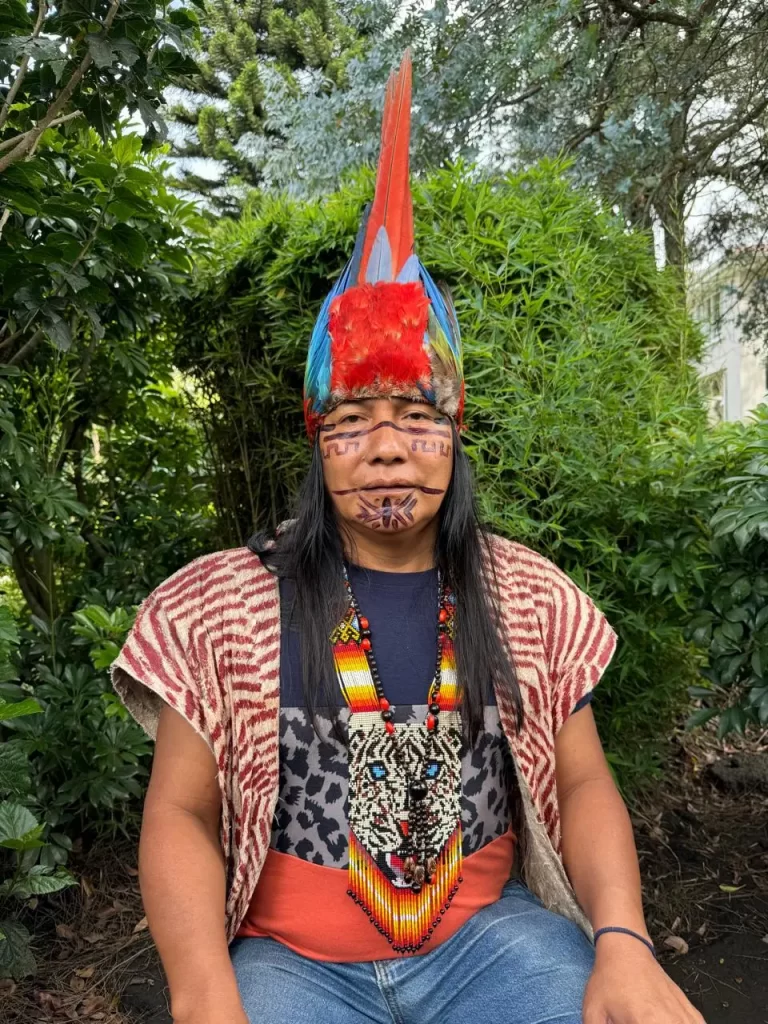 Una persona con cabello largo se sienta al aire libre, lleva un tocado de plumas de colores brillantes, un collar tradicional de cuentas y ropa estampada. Tiene la cara pintada con diseños intrincados. Está rodeada de vegetación, incluidos arbustos y árboles.