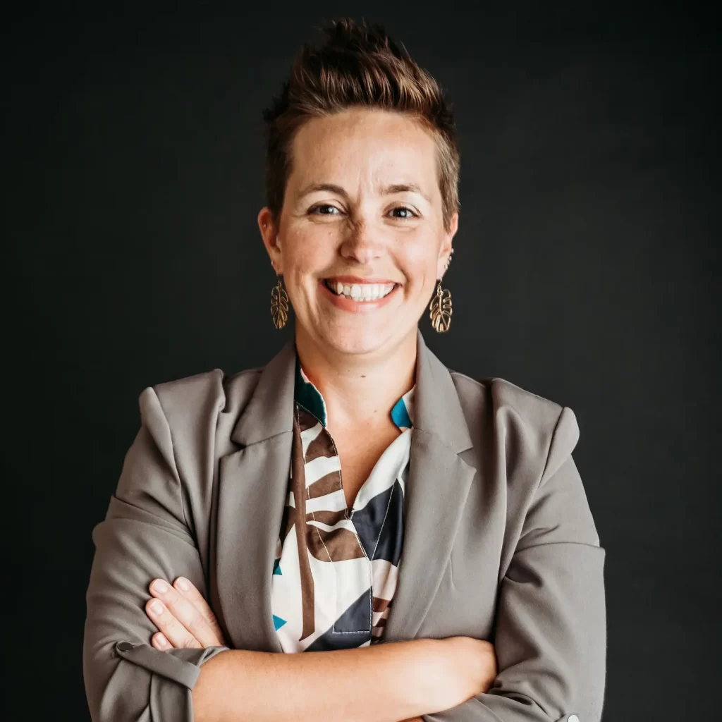 A person with short hair smiles confidently while standing with arms crossed. They are wearing a gray blazer over a patterned shirt and have gold earrings. The background is a plain dark backdrop.