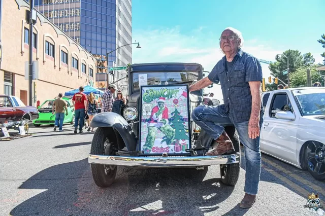 Un hombre mayor se encuentra de pie junto a un coche clásico en la calle, con un pie sobre el parachoques. En el frontal del coche se puede ver un cartel colorido de "El Grinch". Al fondo, bajo un cielo soleado, se ven otros coches clásicos y personas.